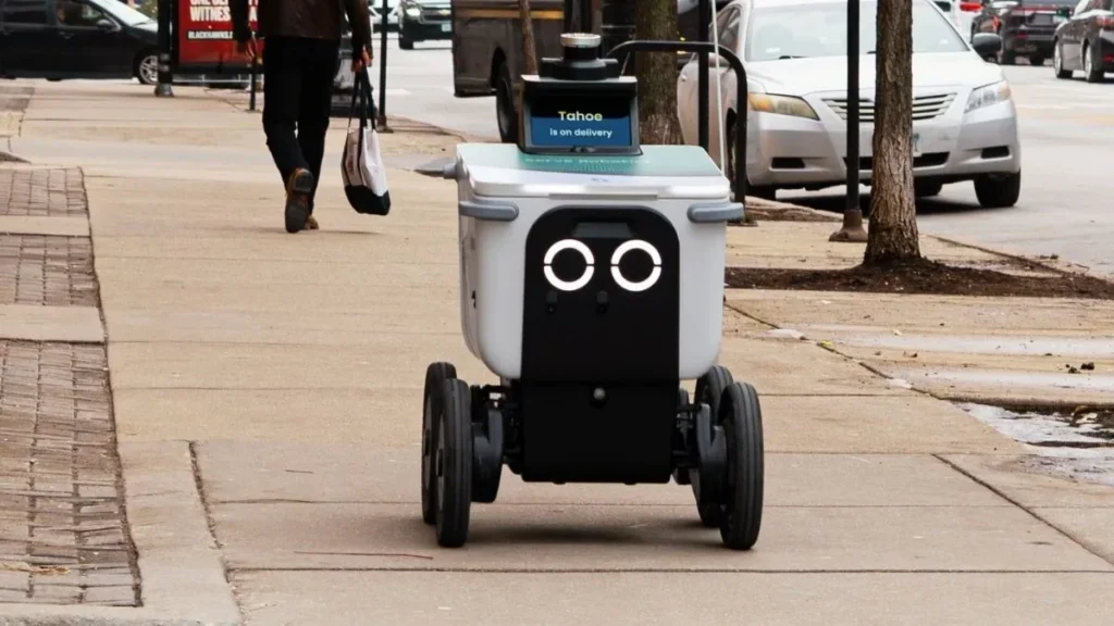 Robô entregador autônomo andando na calçada de uma rua urbana.