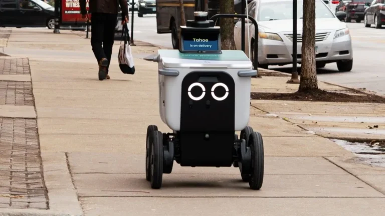 Robô entregador autônomo andando na calçada de uma rua urbana.