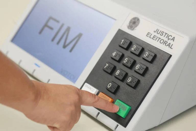 Eleitor pressiona a tecla verde de confirmação de uma urna eletrônica brasileira, com a palavra FIM exibida na tela.